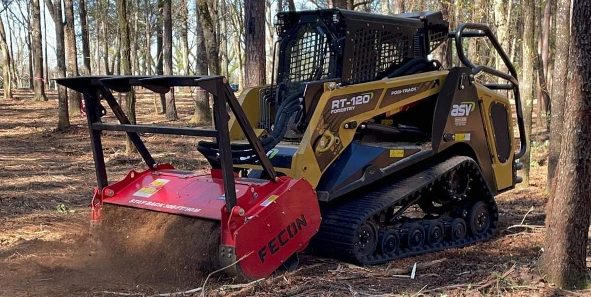 Yellow skid steer with red forestry mulcher attachment clearing brush in a wooded area