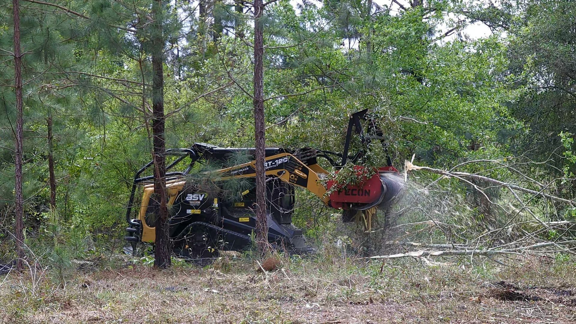 Yellow excavator clearing brush in a wooded area, with branches and debris scattered nearby.