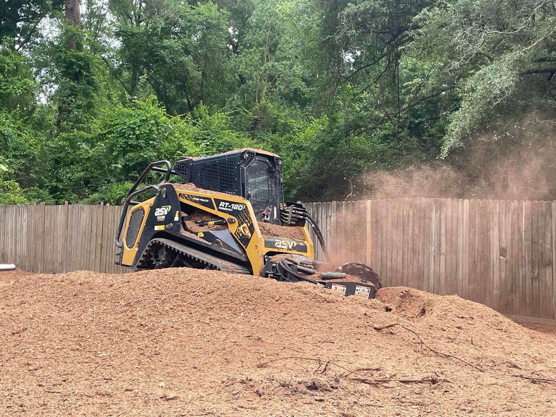 Yellow excavator working on a dirt mound beside a wooden fence and trees
