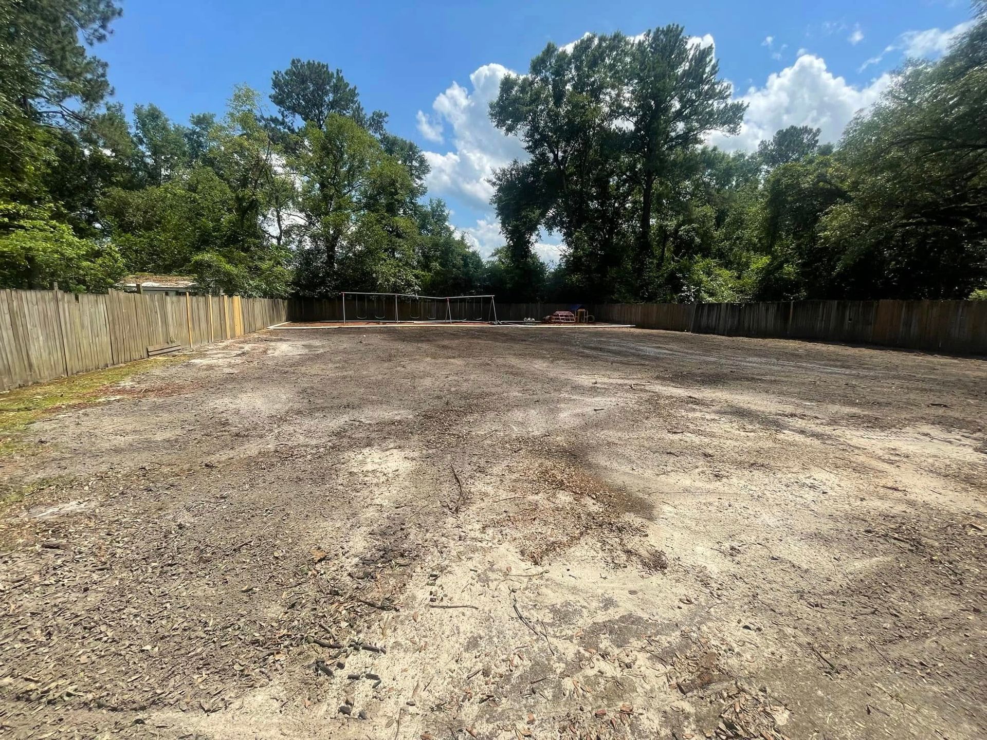 Cleared dirt lot bordered by wooden fences and trees under a bright blue sky with clouds