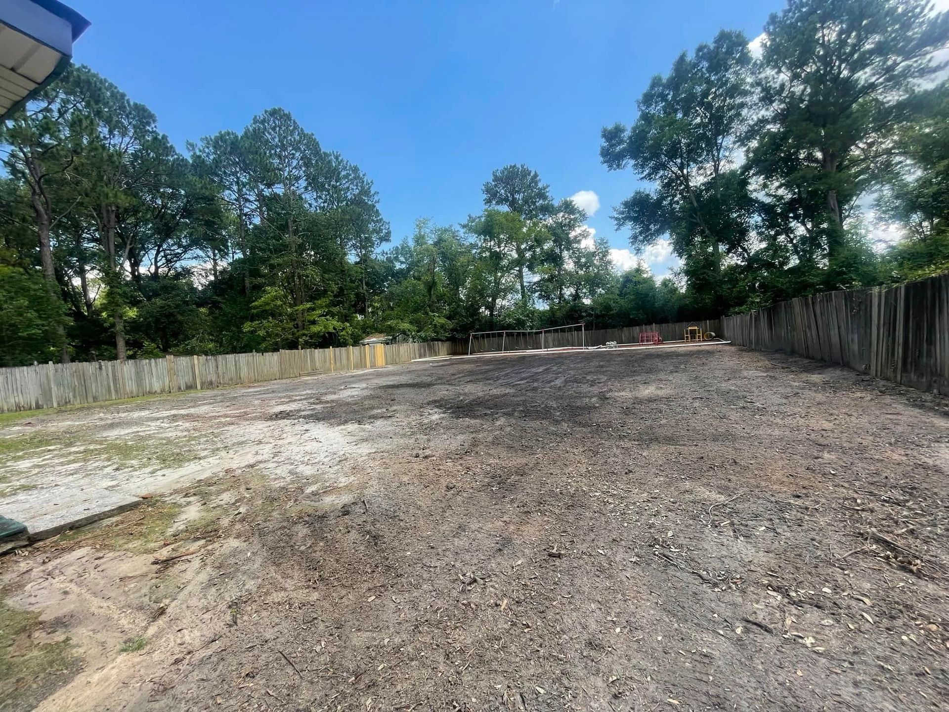 A large cleared dirt lot bordered by wooden fences and trees under a blue sky.