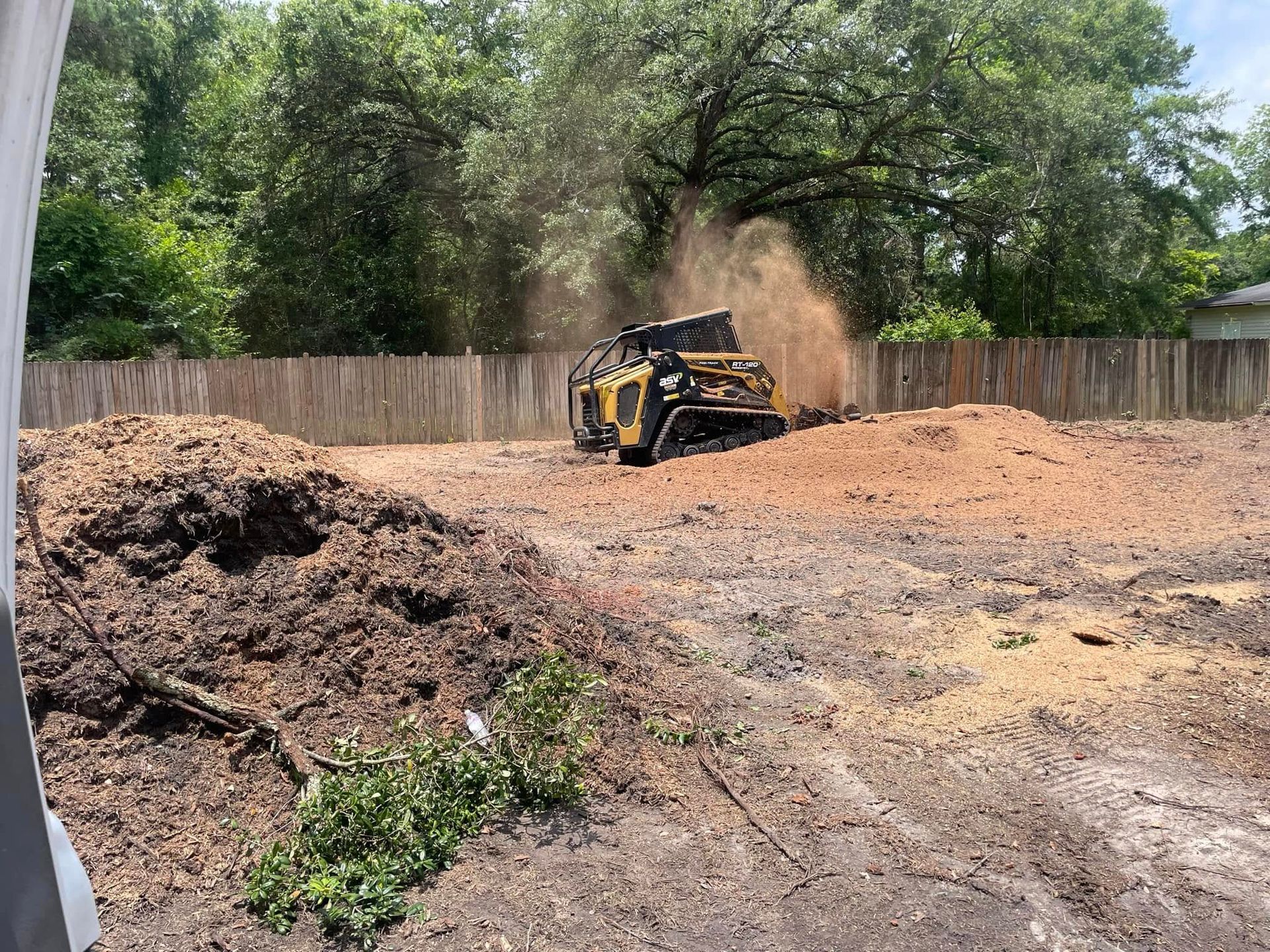 Yellow skid steer moving dirt in a fenced backyard, kicking up dust near trees.