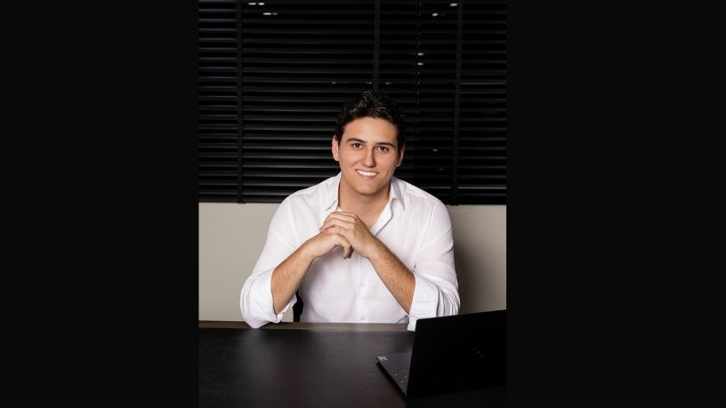 Homem sentado à mesa com as mãos entrelaçadas, vestindo uma camisa branca, em um escritório escuro.