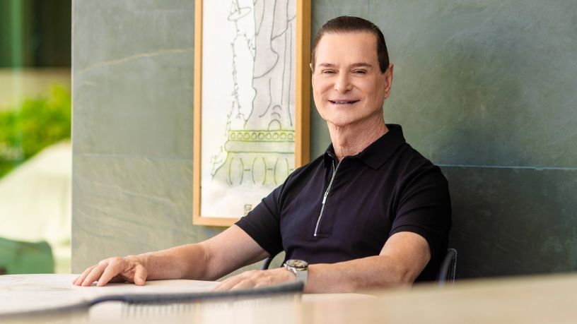 A smiling man in a black polo shirt seated by a green wall and window