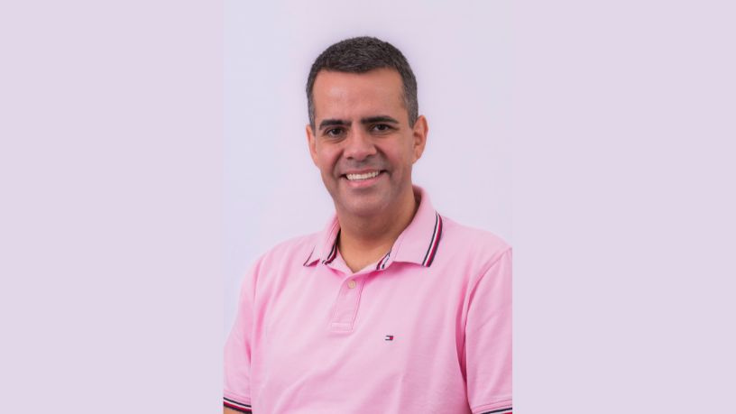 Homem sorridente com camisa polo rosa contra um fundo lilás claro.