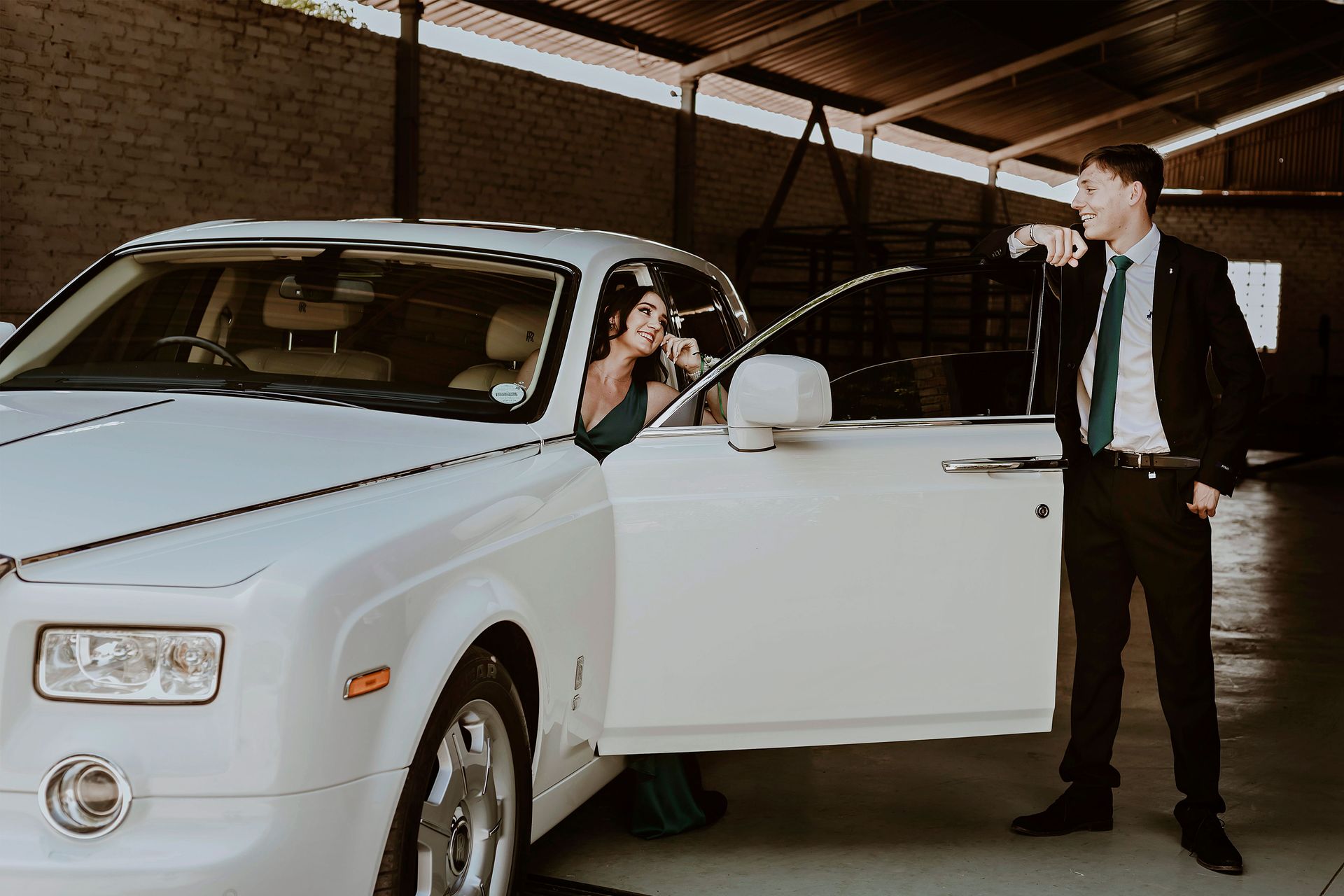 White luxury car in a barn-like garage, with a person standing outside the open door and another inside waving.