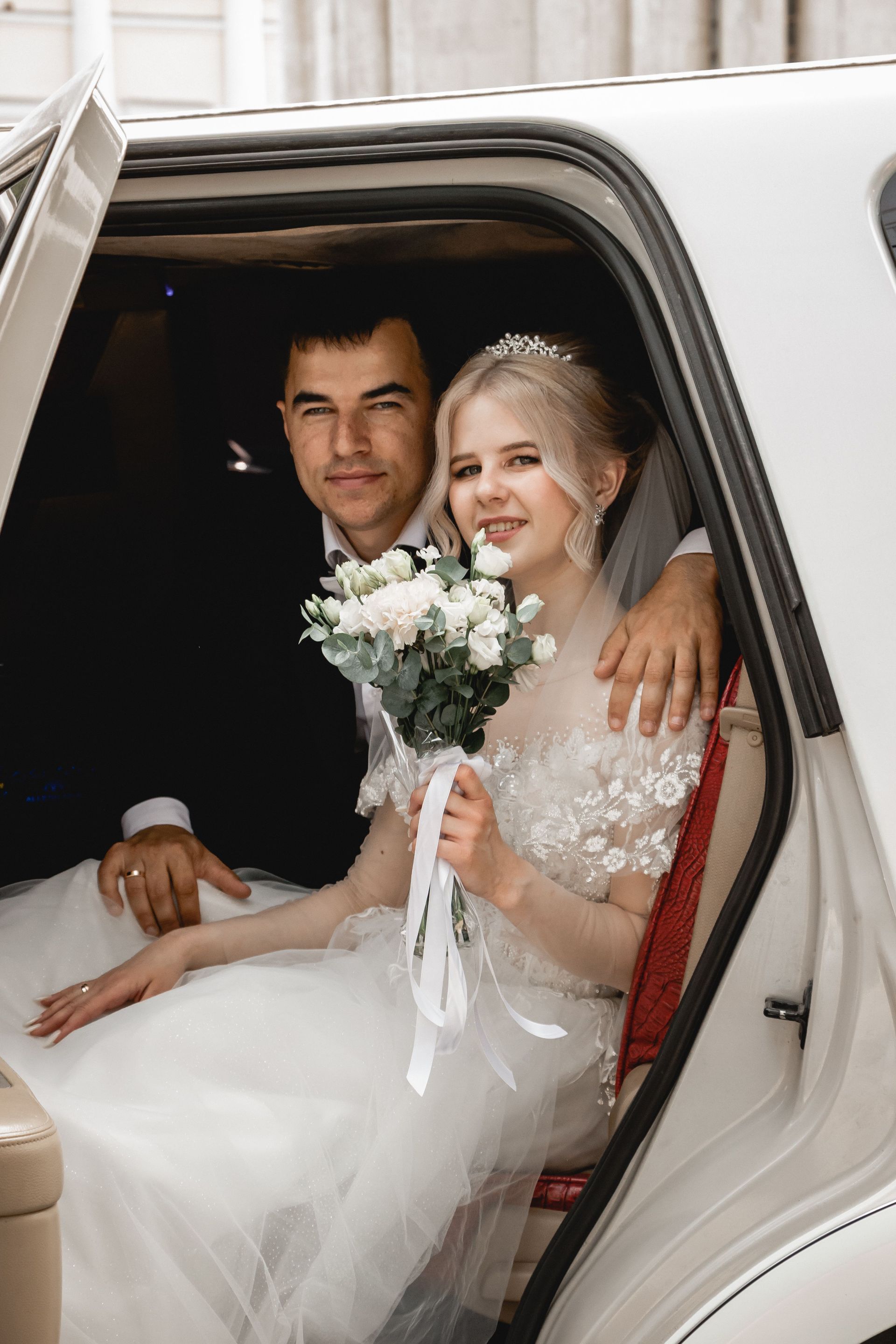 Newlyweds seated in a car, smiling and holding a white bouquet.