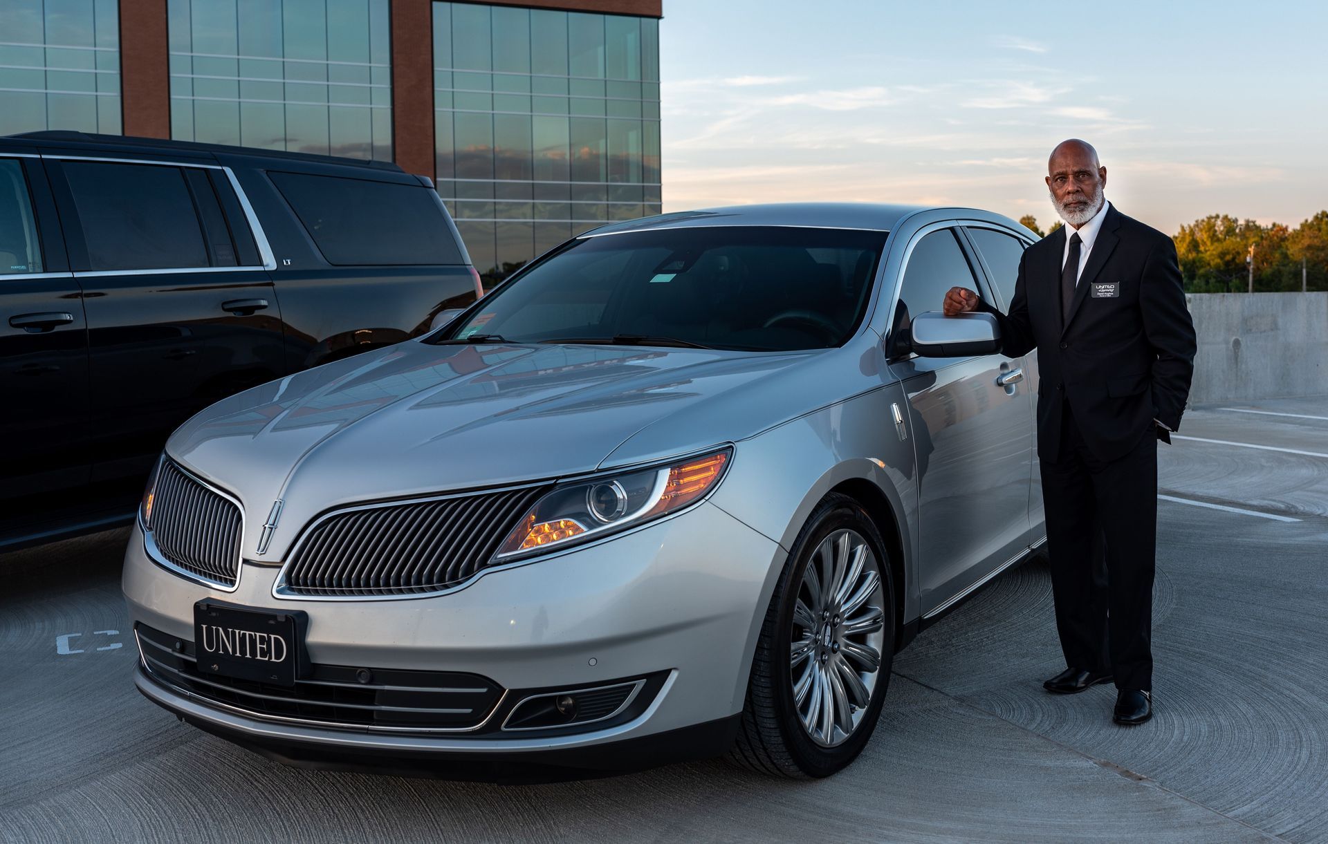 Man in a black suit standing beside a silver luxury sedan outside a modern building.