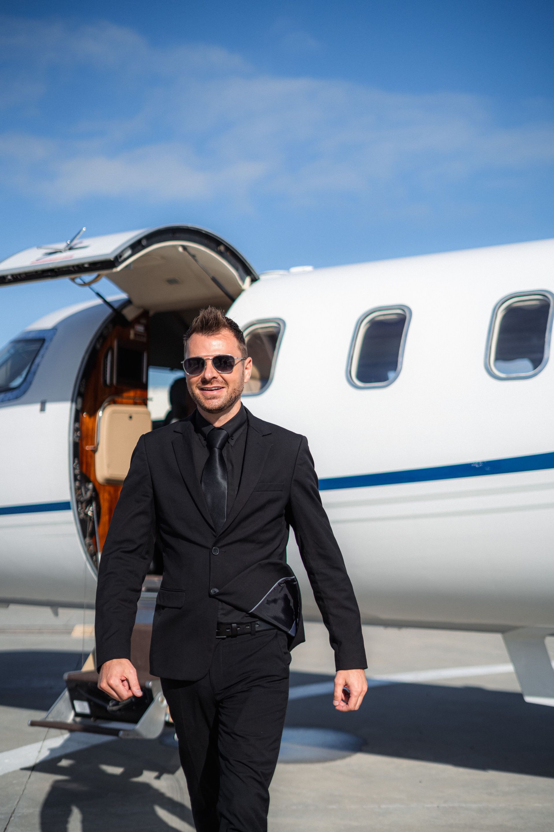 Man in a black suit and sunglasses walking beside a private jet with the door open