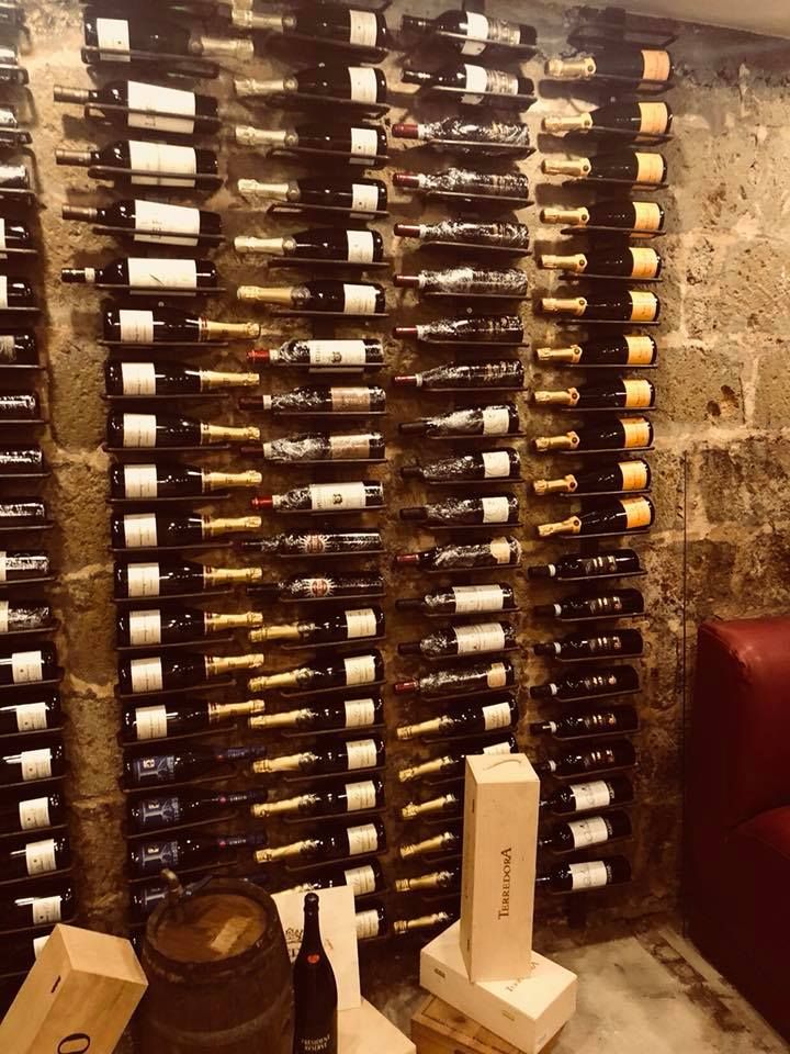Portabottiglie a muro, pieno di bottiglie, accanto a un muro di pietra, con un tavolo di legno e un secchio sottostanti.