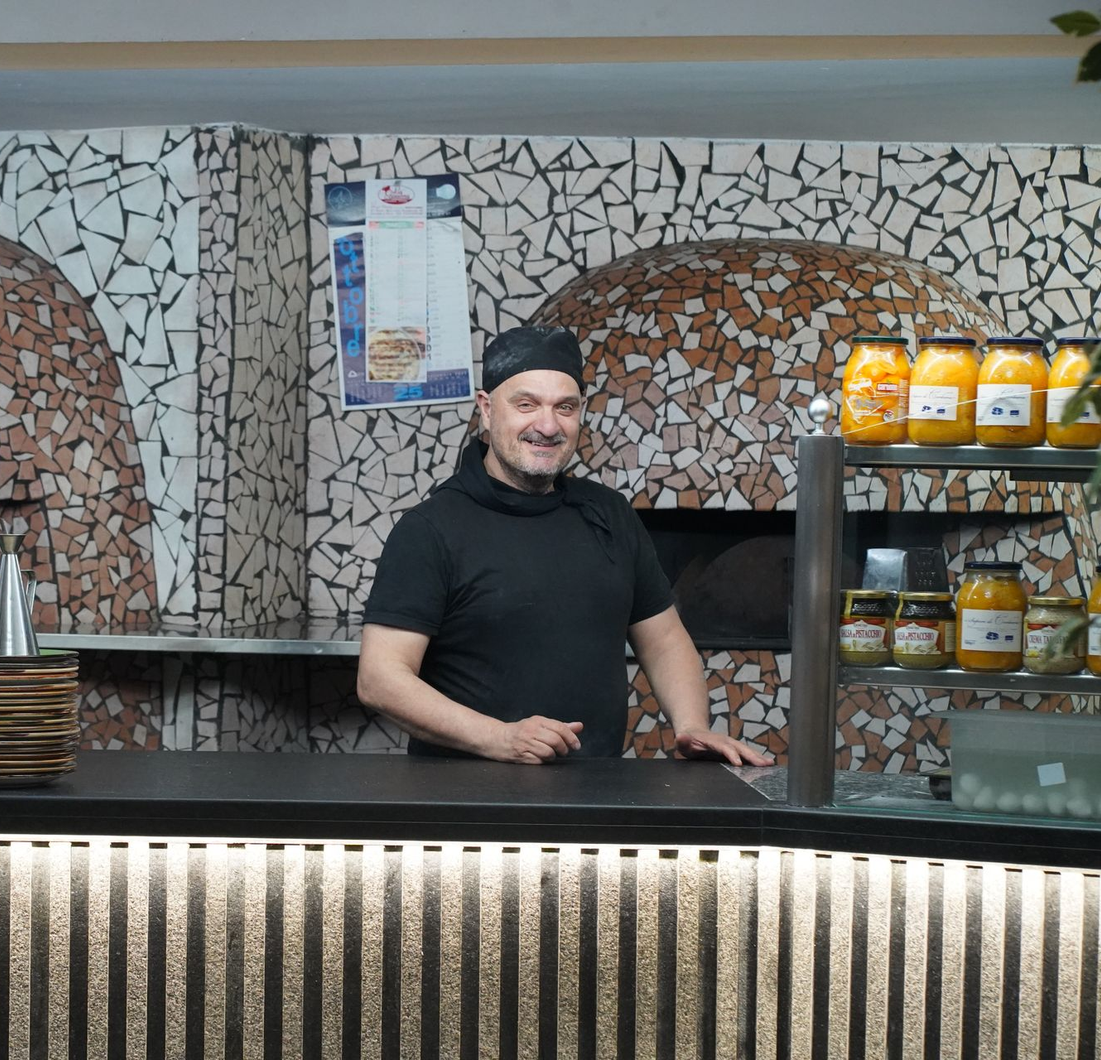 Un uomo in piedi dietro il bancone di un bar, con barattoli di miele e uno sfondo di pareti rivestite di piastrelle a mosaico.