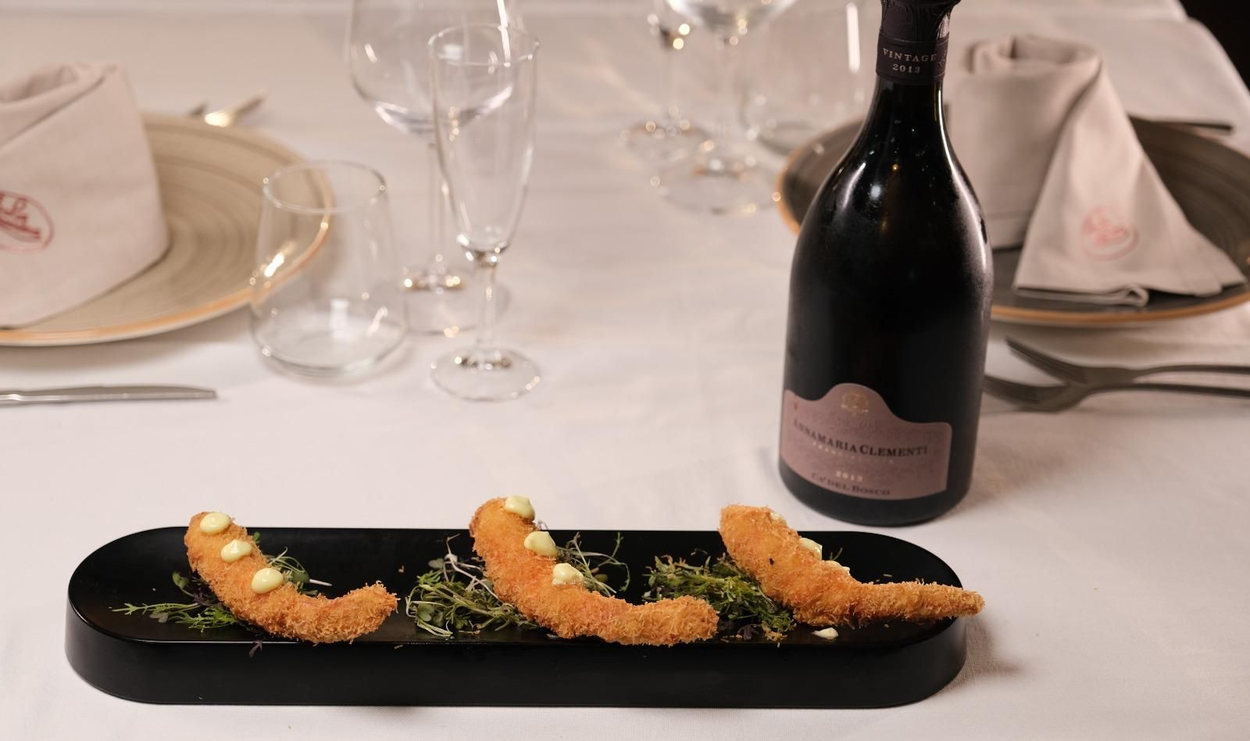 Tre involtini di sushi su un piatto nero, adagiati su un tavolo bianco con bicchieri e una bottiglia di vino.