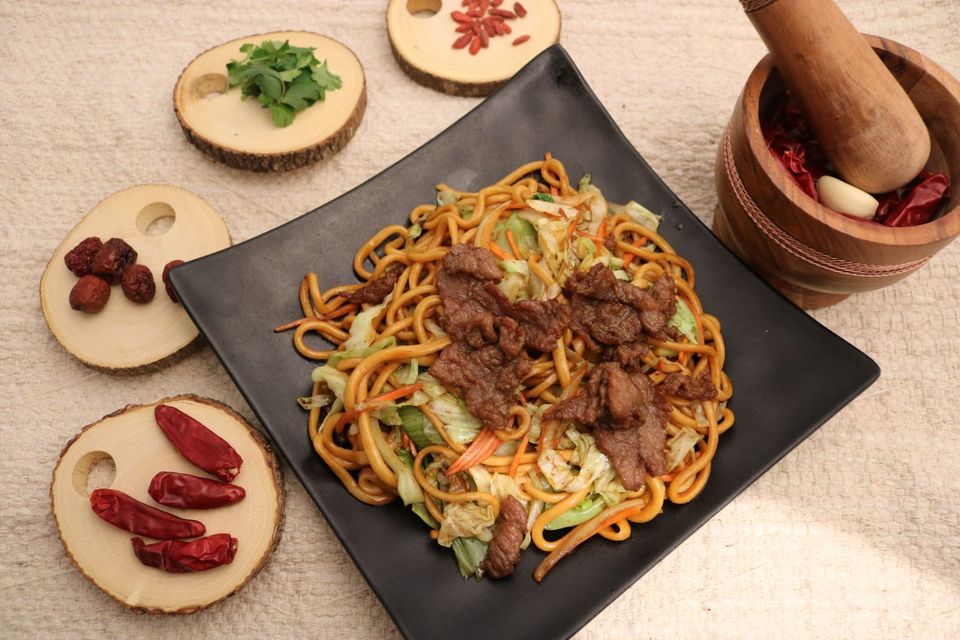 Plated noodles with meat and vegetables, surrounded by small bowls of spices and herbs on a beige table.