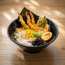 Black ramen bowl with tempura shrimp, seaweed, corn, egg, and vegetables on a wooden table