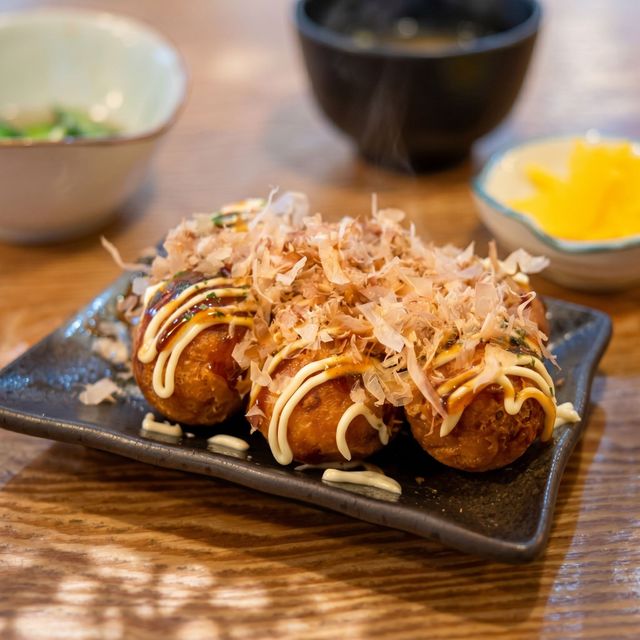 Takoyaki topped with mayonnaise and bonito flakes on a black plate, with soup bowls in the background