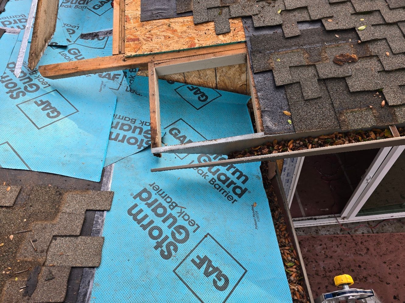 Roof repair with blue underlayment, exposed wood framing, and dark shingles removed around an opening