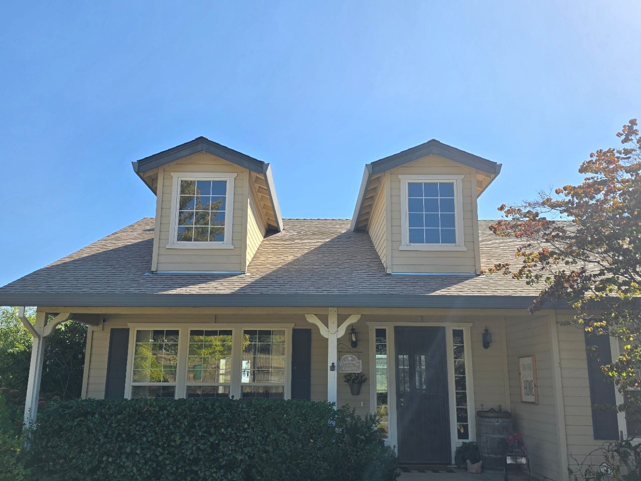 Small house with a gray shingled roof, dormer windows, and a front porch behind a hedge under a blue sky