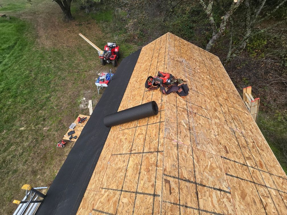Roof under construction with black underlayment, tools, and workers on plywood sheathing.