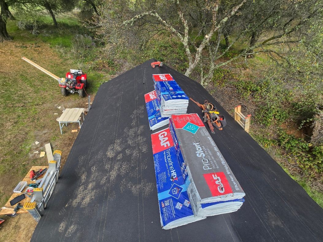 A tractor carries stacks of blue and red roofing materials on a steep roof in a wooded area.