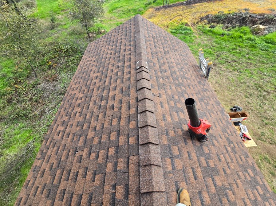 Roof ridge with brown shingles and a black vent pipe, surrounded by green grass and trees.
