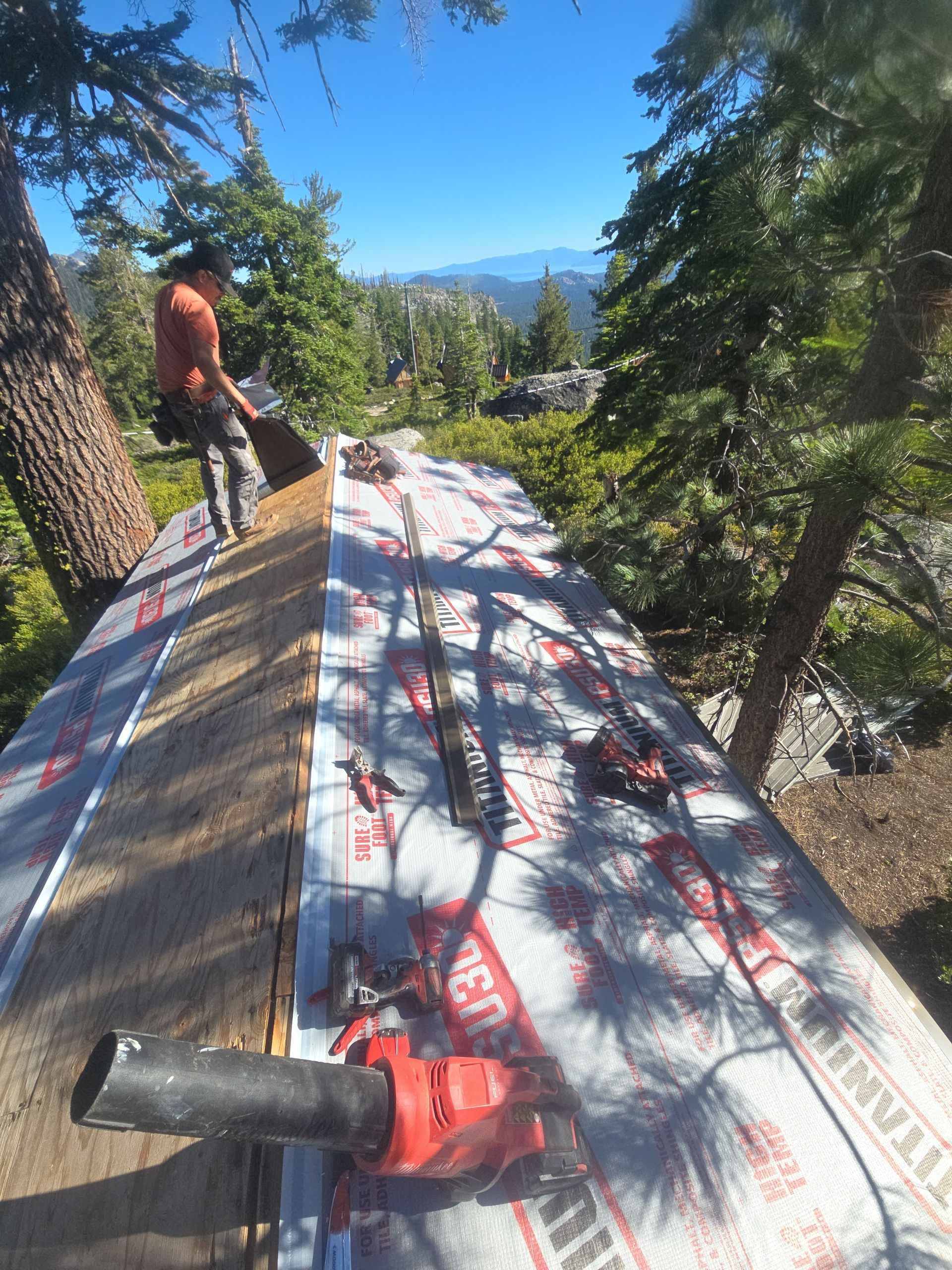 Worker installing roofing underlayment on a steep roof among tall pine trees, with mountains in the background