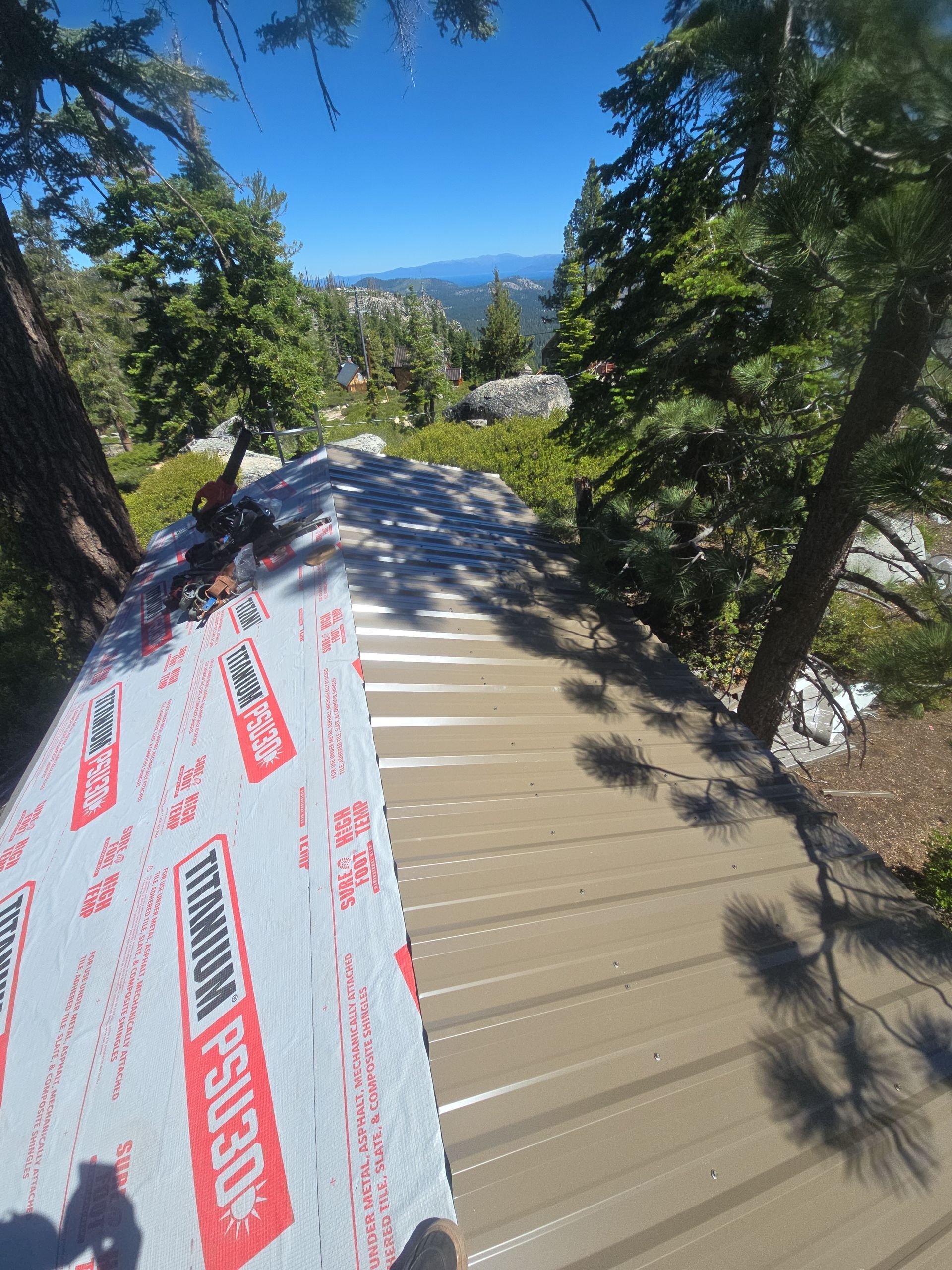 Rooftop under repair with white roofing underlayment, surrounded by tall trees and mountain views