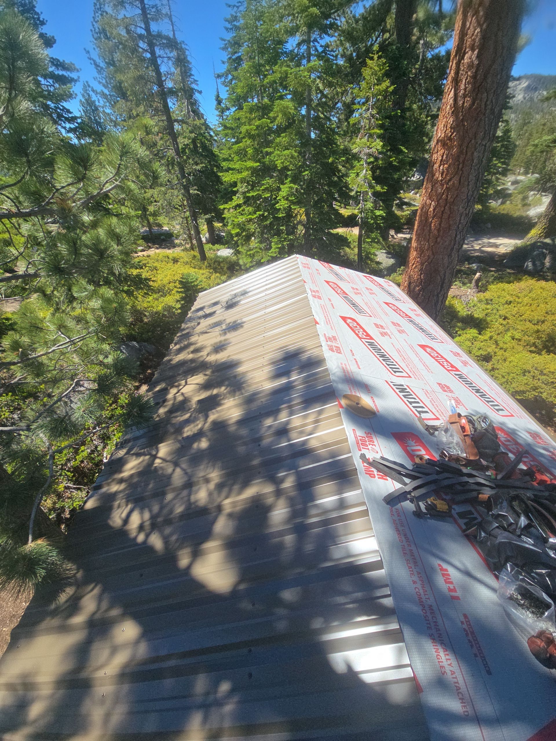 Roof under construction in a wooded area, with gray shingles and white underlayment covered in red text.