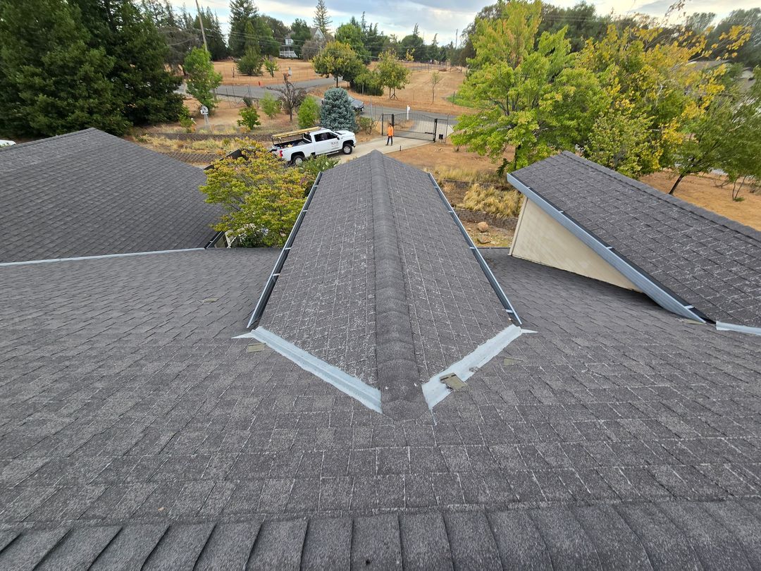 View from a shingled roof over a neighborhood with trees, parked cars, and dry grass in the distance