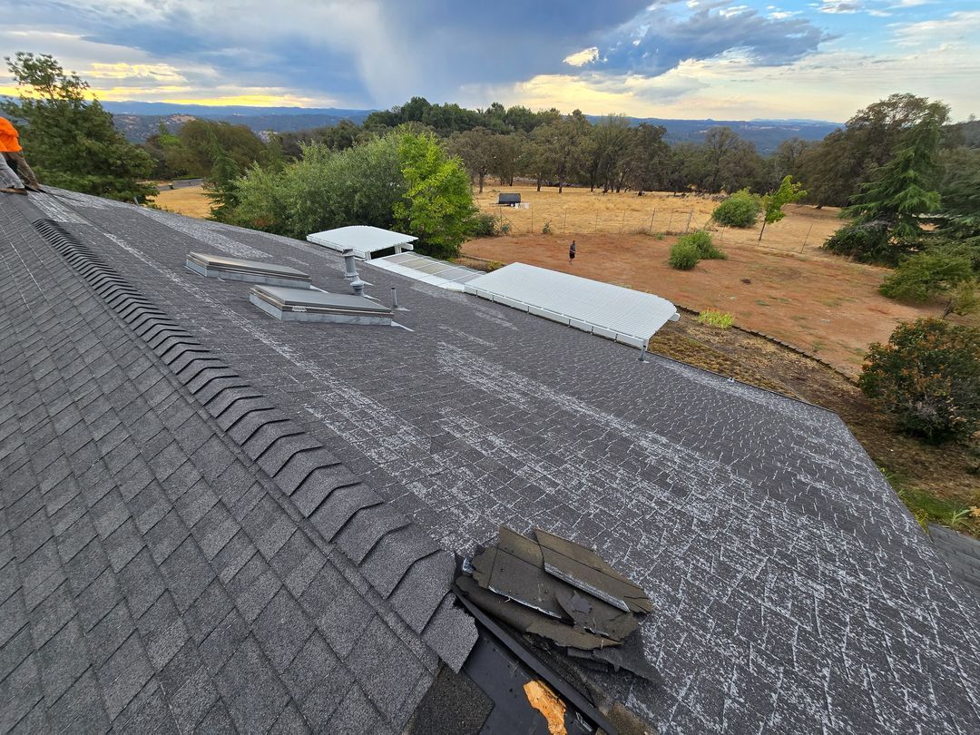 Rooftop under repair with dark shingles, white underlayment, and a view of a dry landscape at sunset