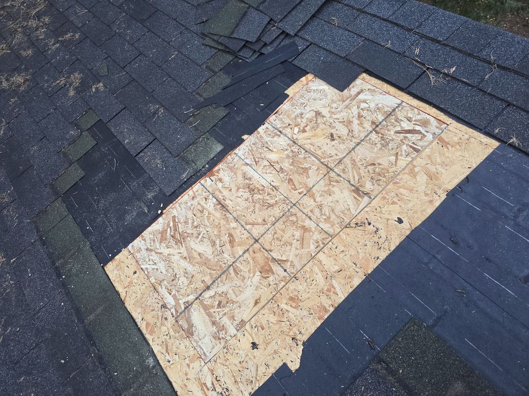 Damaged roof with a light-colored plywood patch surrounded by dark shingles.