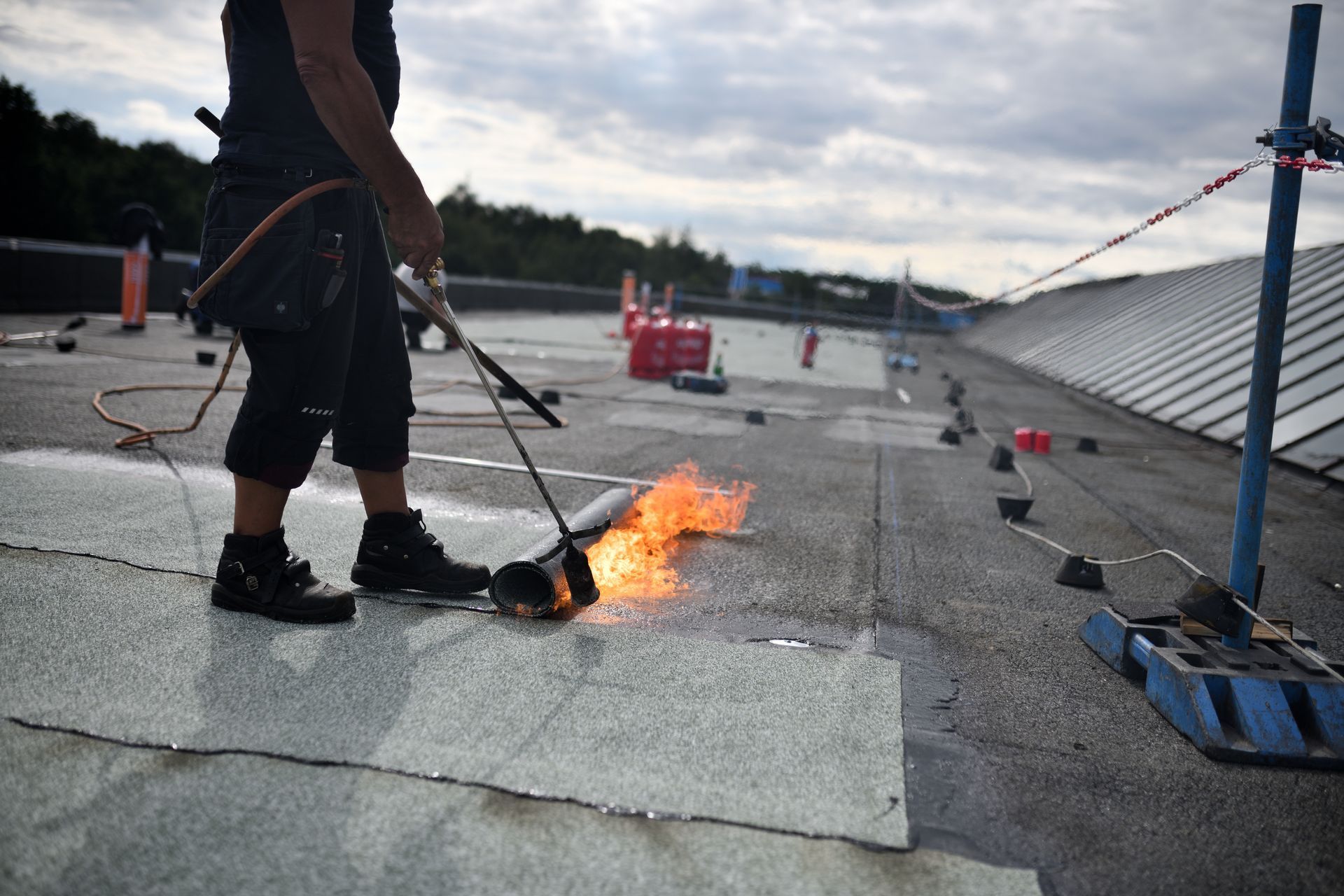 Worker using a torch to burn roofing material on a flat roof with tools and safety cones nearby