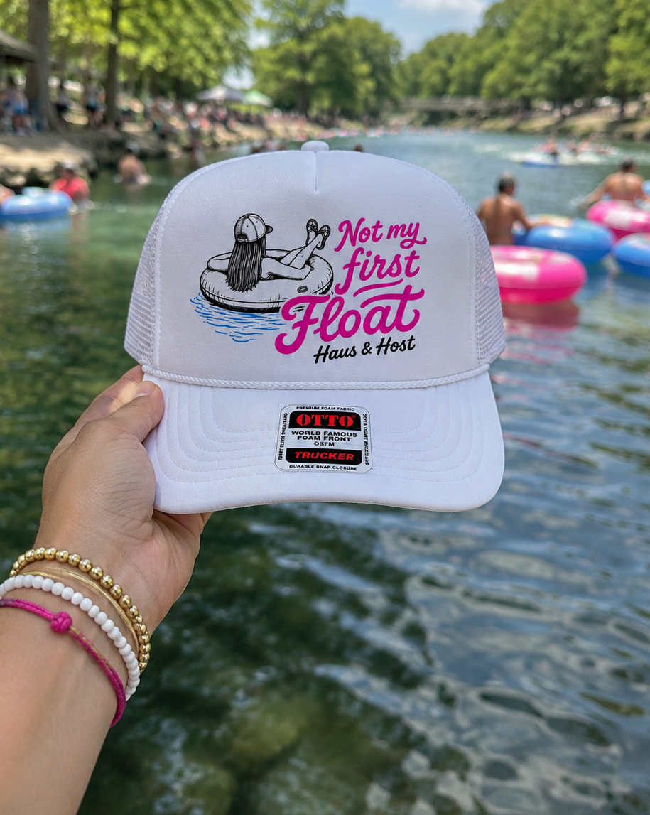 Hand holding a white cap reading “Let me first Float” by a river with colorful floating tubes in the background