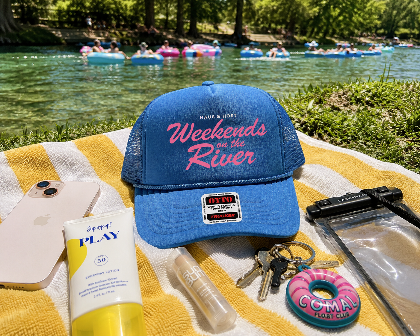 Blue “Weekends on the River” cap on a towel by a river, with sunscreen and keys in the foreground.