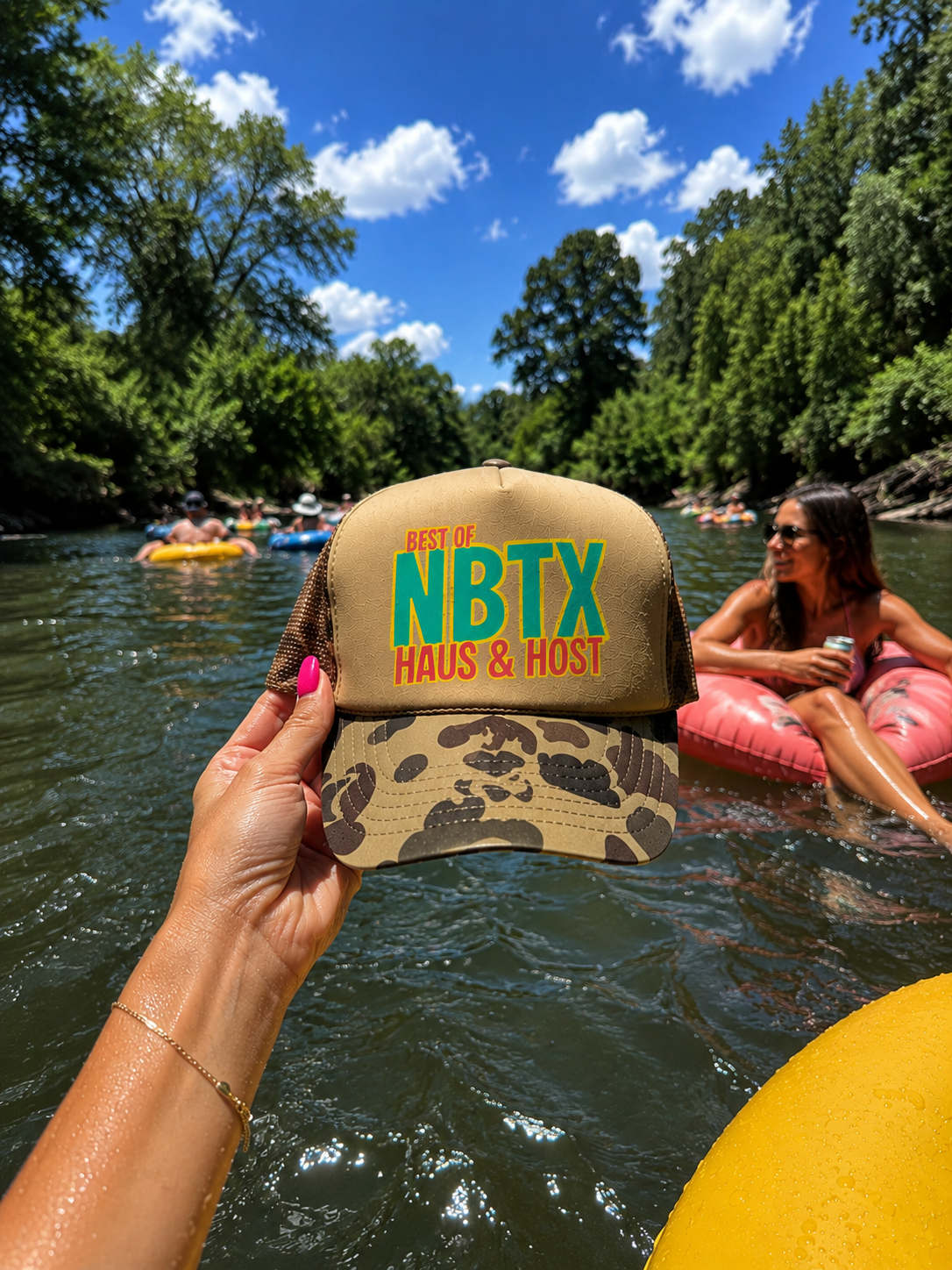 Hands holding camo cap reading “NBTX House of Ast” on a river float with people and trees under a blue sky