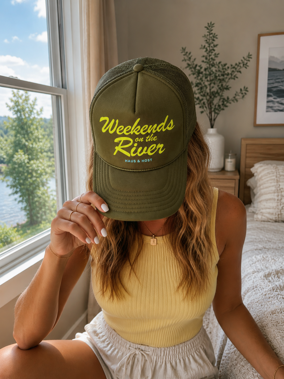 Person indoors by a window, holding up an olive cap with yellow “Weekends River” text.