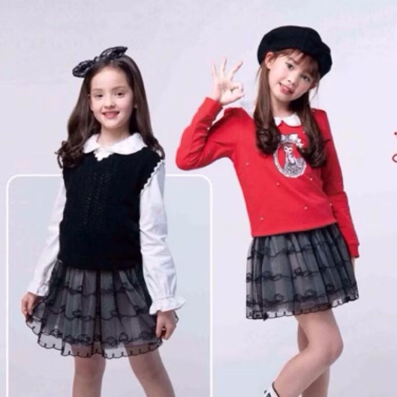 兩個穿著相配裙子的小女孩，一個穿著毛衣背心，另一個穿著紅襯衫，對著鏡頭微笑。