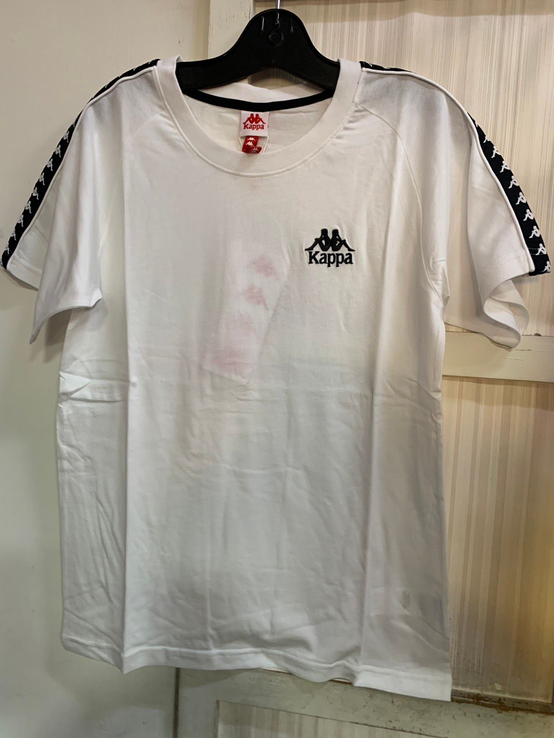 白色 Kappa T 卹，肩部有黑色標誌和條紋細節，掛在黑色衣架上。