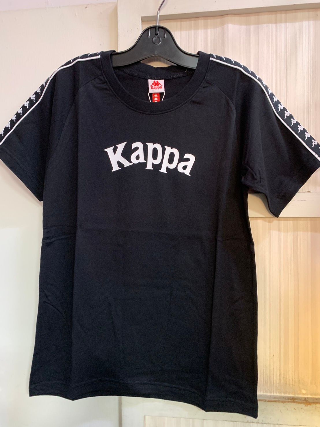 黑色 Kappa T 卹，帶有白色標誌和條紋袖子細節，掛在黑色衣架上。