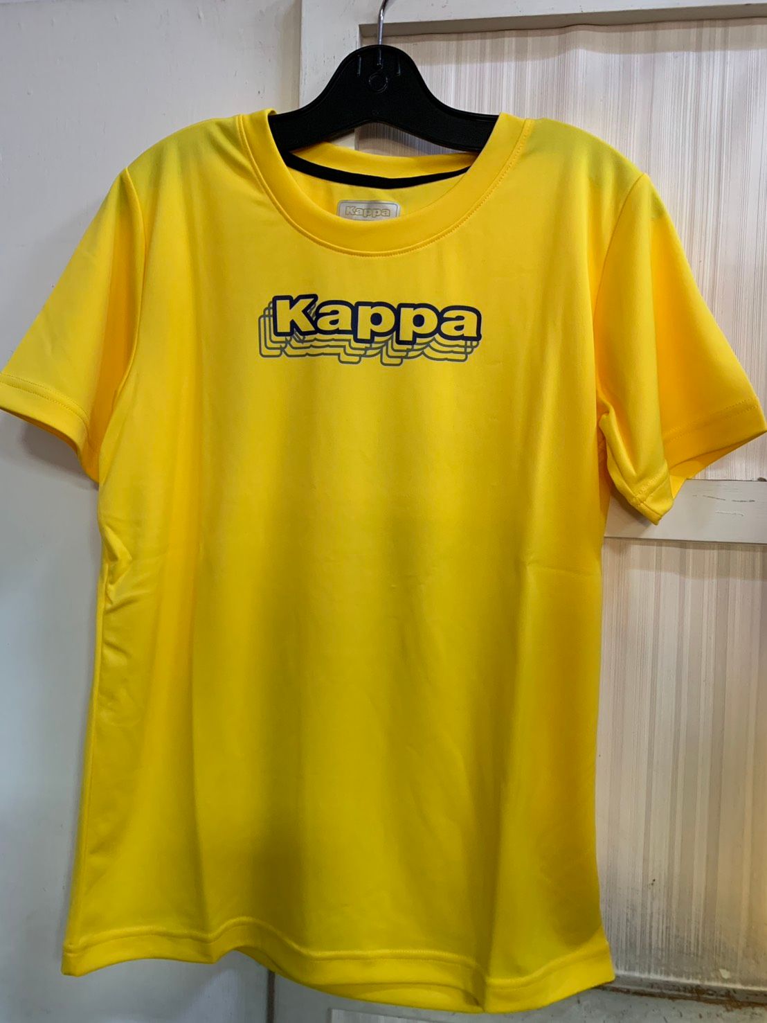 黃色的 Kappa T 卹，帶有黑色和黃色的標誌，掛在黑色的衣架上。