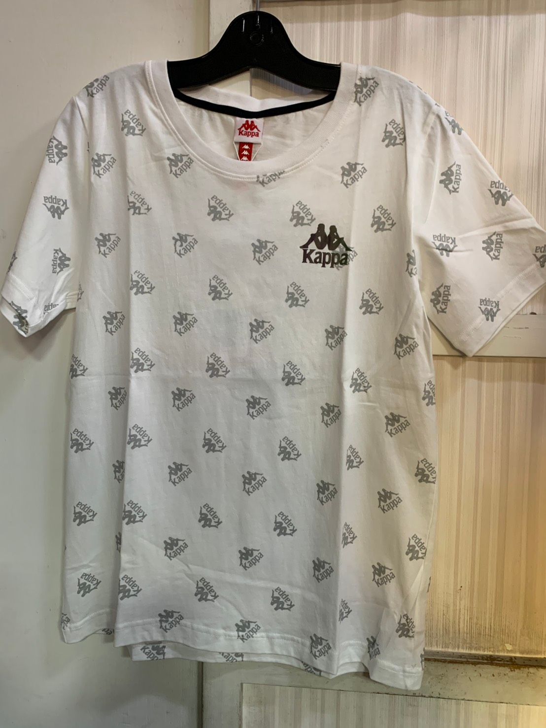 白色 Kappa T 卹，帶有重複的灰色 Kappa 標誌圖案、黑色衣領和胸前的小標誌。