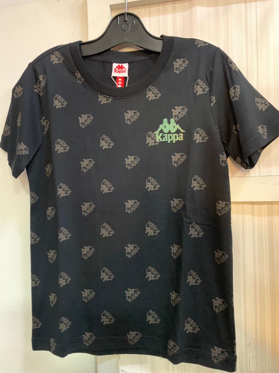 白色 Kappa T 卹，帶有重複的灰色 Kappa 標誌圖案、黑色衣領和胸前的小標誌。