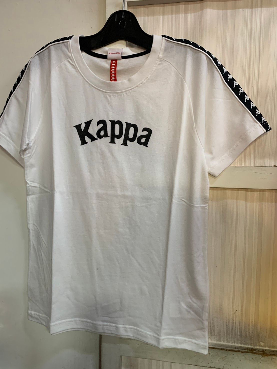 黑色 Kappa T 卹，帶有白色標誌和條紋袖子細節，掛在黑色衣架上。