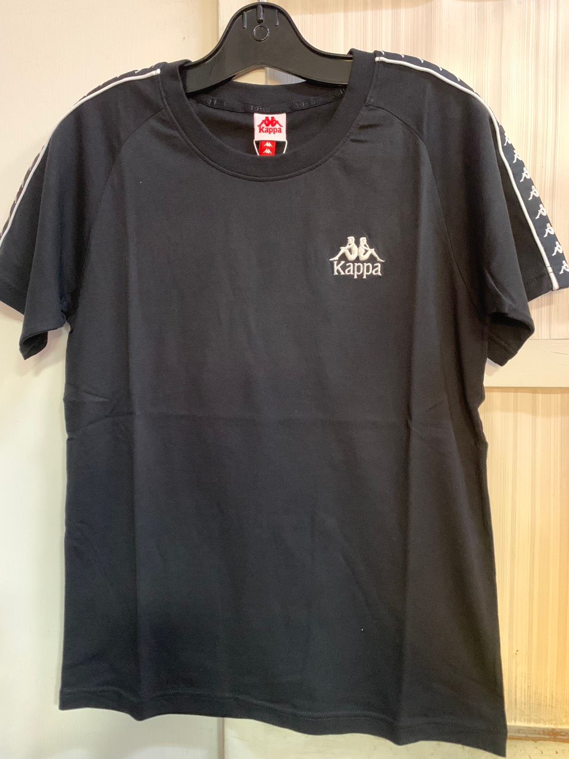 白色 Kappa T 卹，肩部有黑色標誌和條紋細節，掛在黑色衣架上。