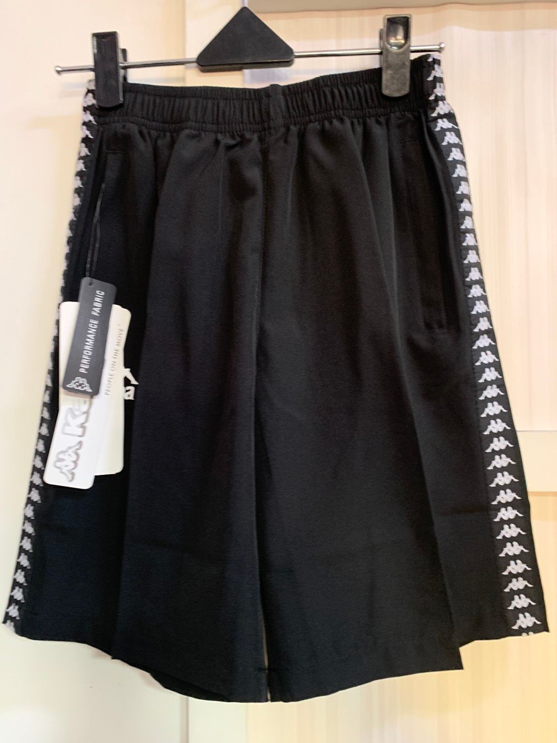 黑色 Kappa 短褲，兩側有白色標誌膠帶，掛在黑色衣架上。