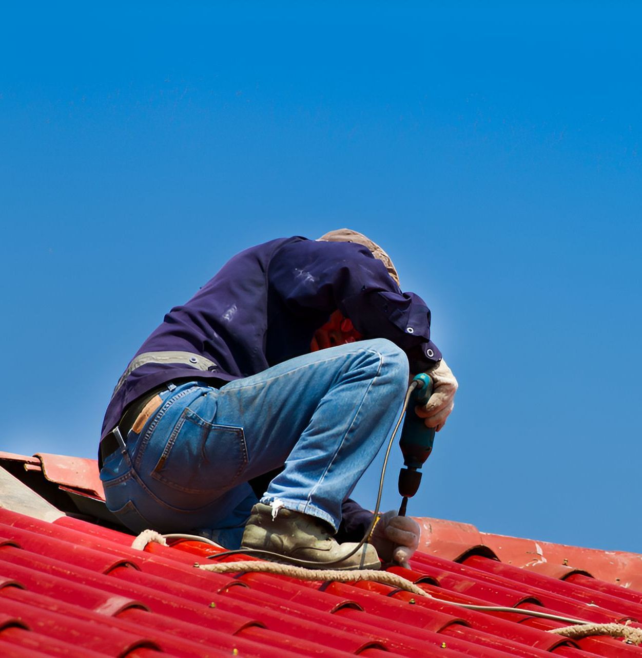 A Man Is Kneeling On A Roof Using A Drill — Roof & Guttering Solutions In Saratoga, NSW