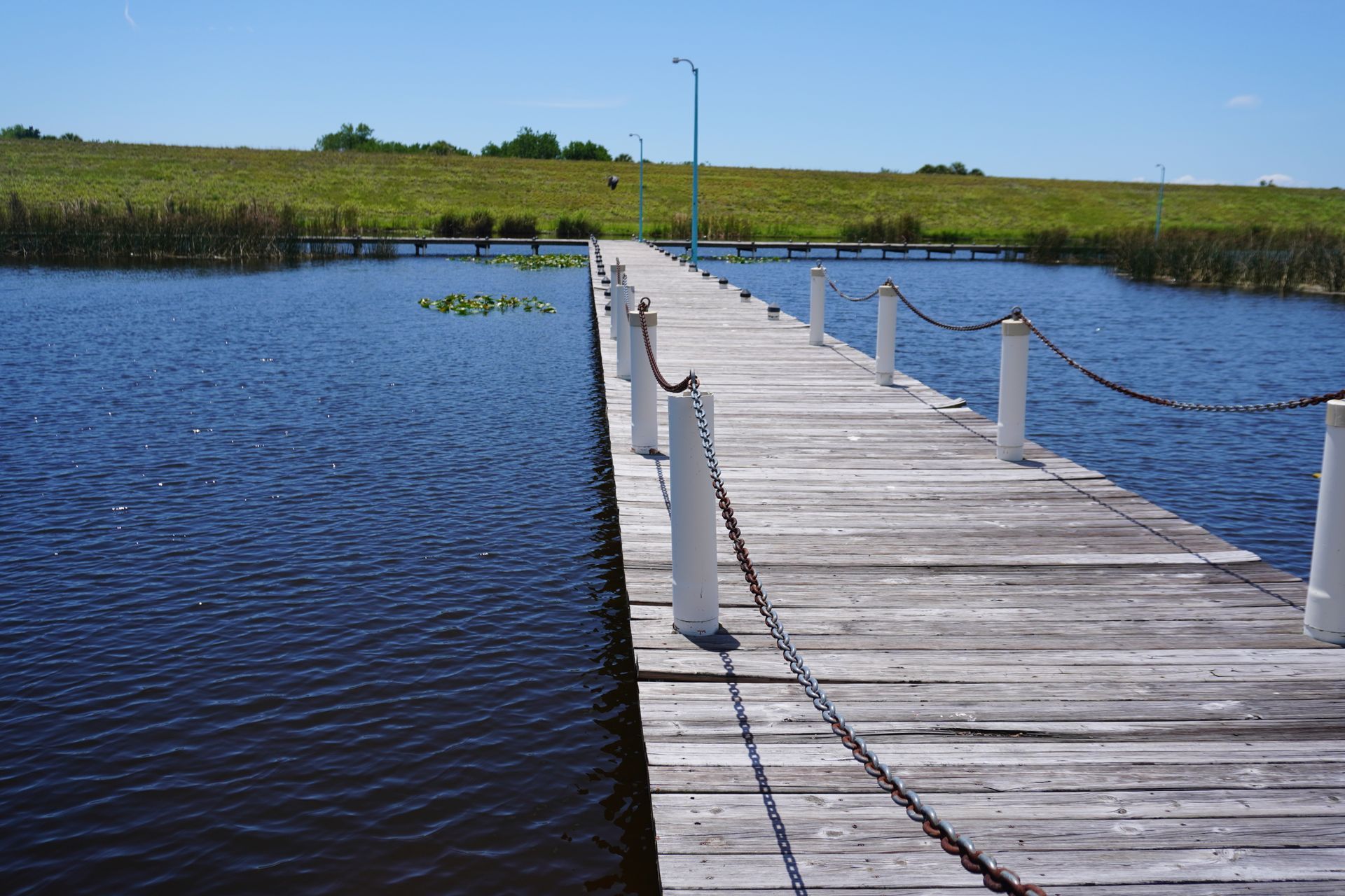Wooden dock extending across calm blue water toward a grassy shoreline under a clear sky