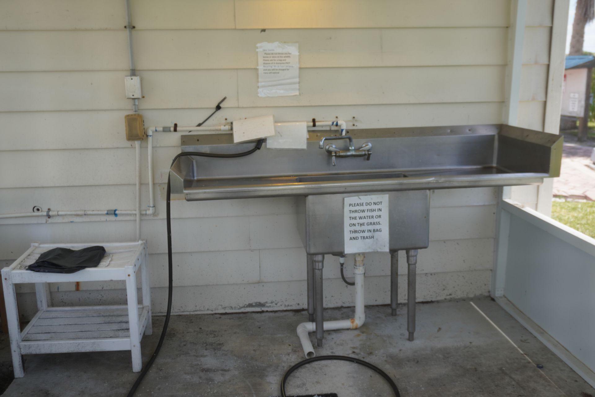 Stainless steel utility sink in a tiled utility room, with a hose and small white side table.