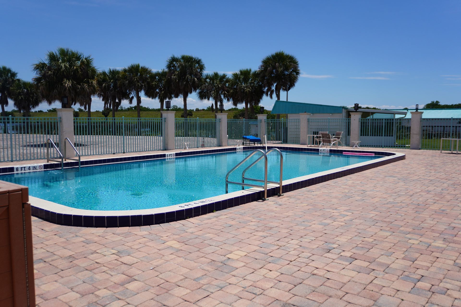 Outdoor swimming pool beside palm trees and a brick patio, with a view of the water beyond.