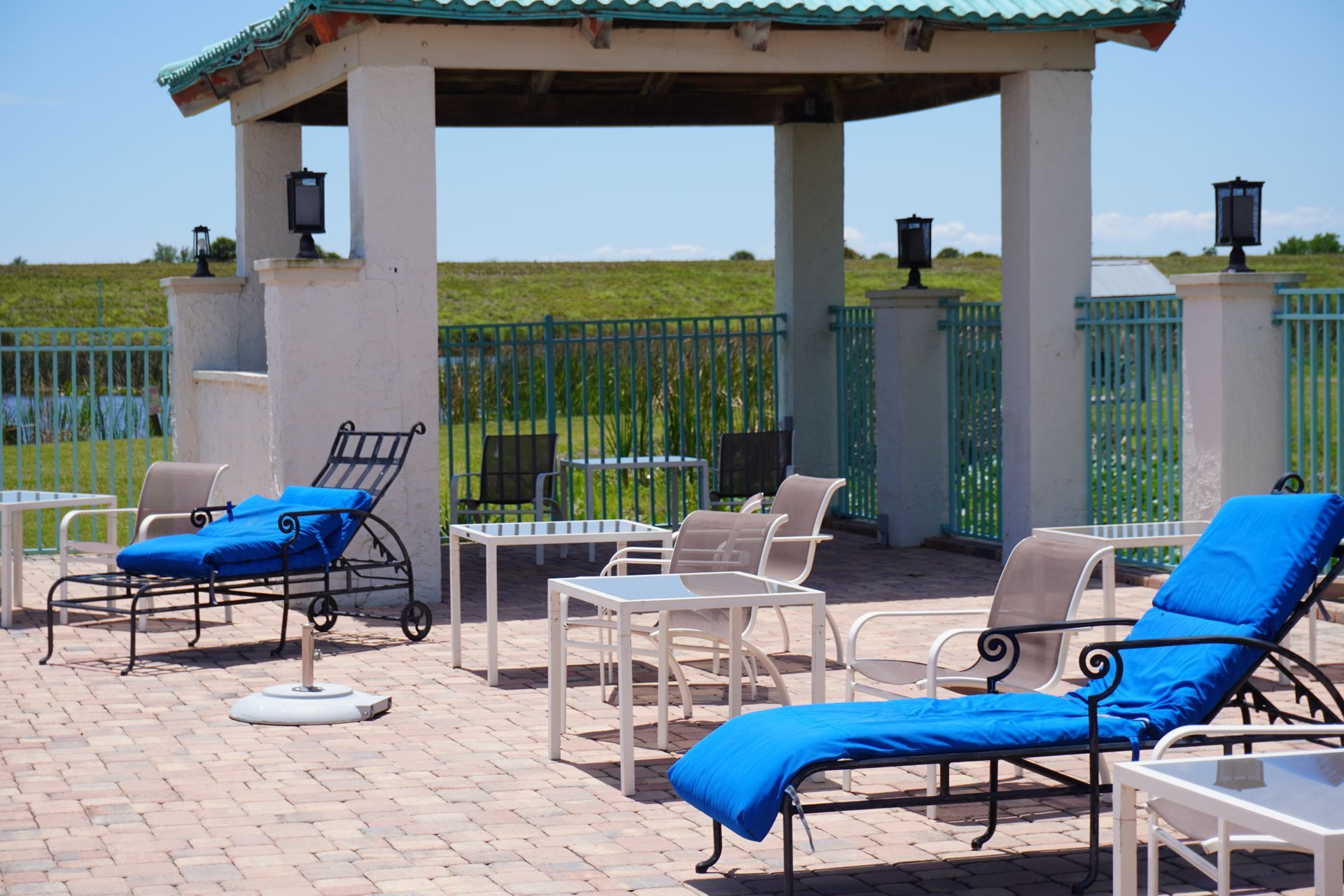 Poolside lounge chairs under a shaded pavilion beside a grassy waterfront fence