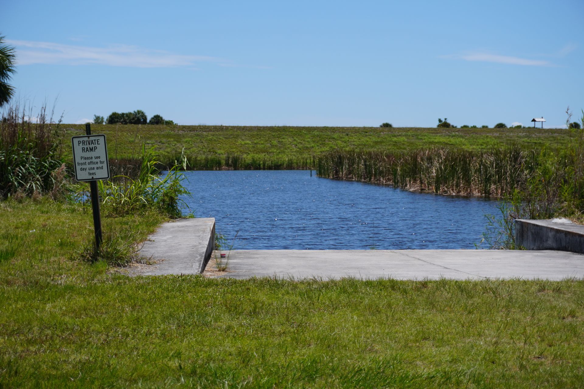 Small concrete boat ramp leading to a blue water channel with tall reeds and open grassy fields under a clear sky